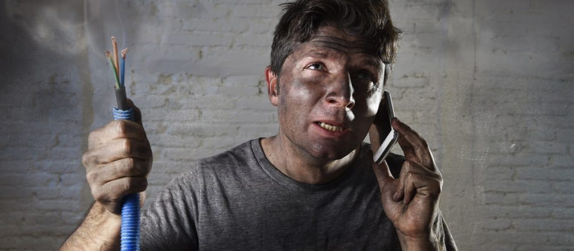 Young man with a dirty, burnt face holding a smoking electrical cable after a DIY mishap, desperately calling for an electrician in New Orleans for help.